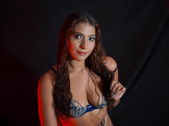 MiaBianchi with wet curly hair, wearing a blue lace bra, looking at the camera, holding her bra strap, red light on her right side, smiling.