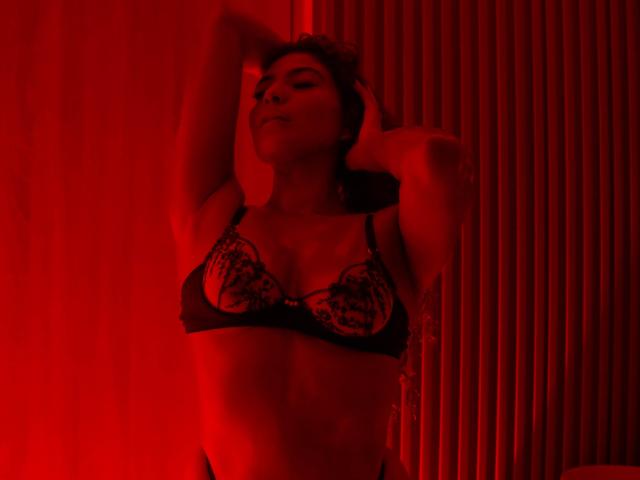 MiaBianchi with hands behind head, wearing black lace bra, red-lit, showing small tits, slightly open mouth, relaxed, confident, medium skin, dark hair, sexy, seductive pose.