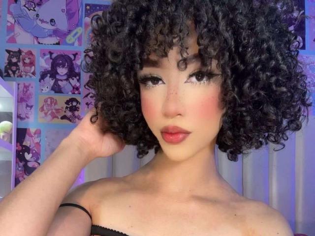 AlissLovely with curly black hair, pink blush, and red lipstick, touching her head with a black strap. Asian face, thick eyelashes, and soft pink cheeks. Topless, showing collarbones.