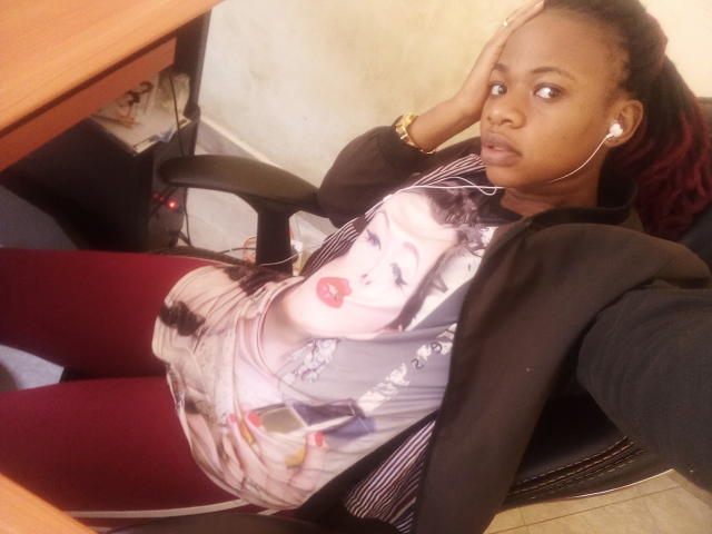 CiaraMary with dark skin, red braids, wearing a black jacket, white shirt with Marilyn Monroe print, red leggings, earbuds, black chair, wooden table, and a wristwatch, looking at camera.