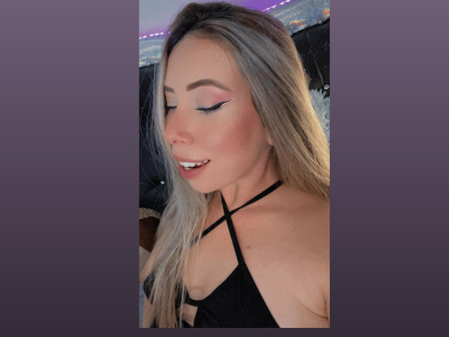 JennyLawry with blonde hair, black strappy top, eyeliner, and pink eyeshadow, showing her upper body and smiling with her eyes closed. Boobs visible, she's cute and sexy.
