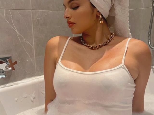 NovaDixon con una canottiera bianca bagnata, capezzoli visibili, grandi seni, pelle abbronzata, un asciugamano in testa, una catena d'oro e orecchini d'oro, seduta in una vasca da bagno piena di bolle..