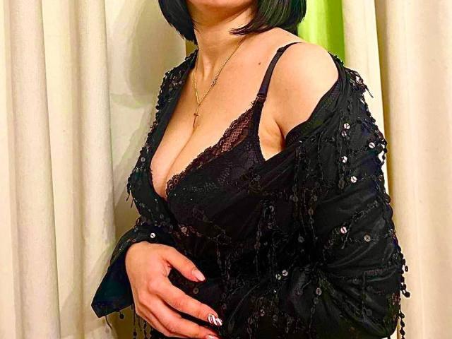 Janel has big tits in a black lace bra, wearing a sequined black shawl, with a short black bob and a gold necklace, standing against curtains.