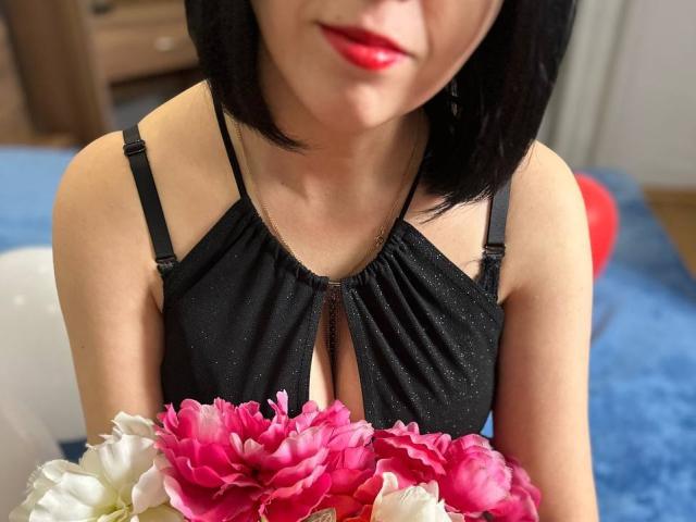 Janel with black bob, red lipstick, black sparkly dress, holding pink and white flowers, showing cleavage, sitting on blue carpet.
