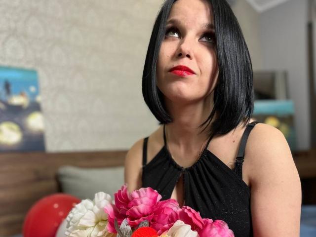 Janel with black bob, red lipstick, black halter dress, holding pink and white flower bouquet, looking up with slight smile, medium-sized tits, fair skin, indoor setting.