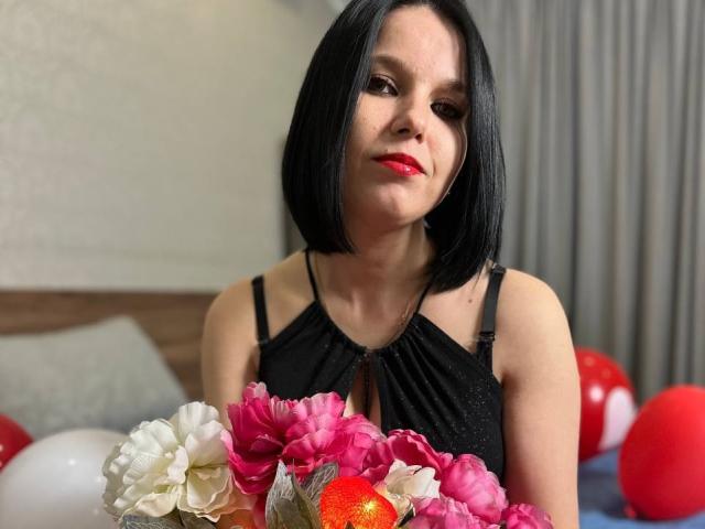Janel with black bob, red lipstick, black dress, holding pink and white flowers, red and white balloons, and a shiny orange fruit, looking slightly to the side.