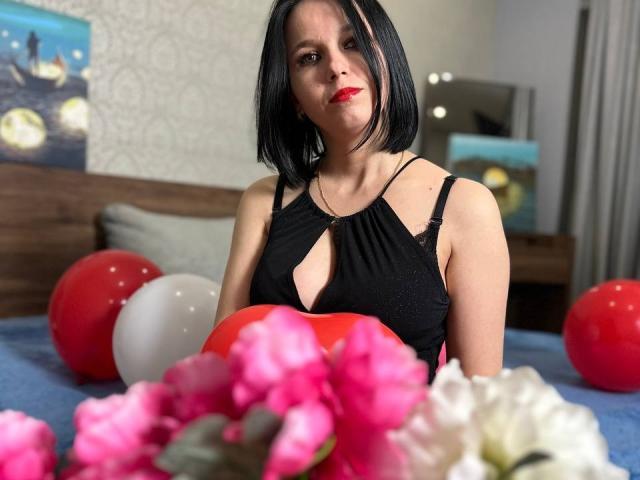 Janel with black bob, red lipstick, wearing black dress with keyhole, sitting on bed with red, white balloons and pink flowers, looking at camera with slight smile.