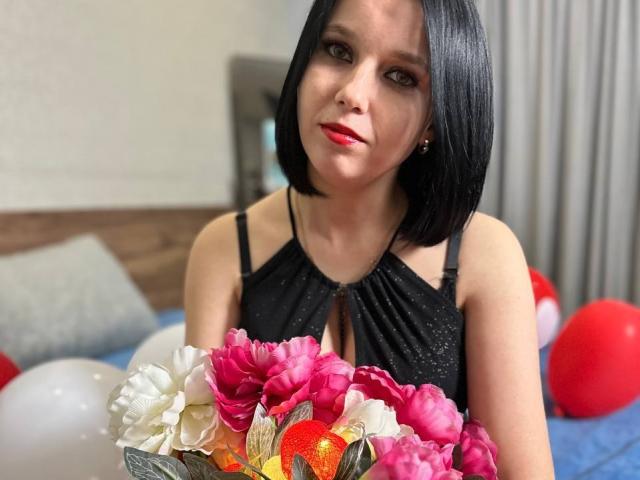 Janel with black bob, red lipstick, black halter dress, holding pink and white flower bouquet, looking at camera. Red and white balloons in background.