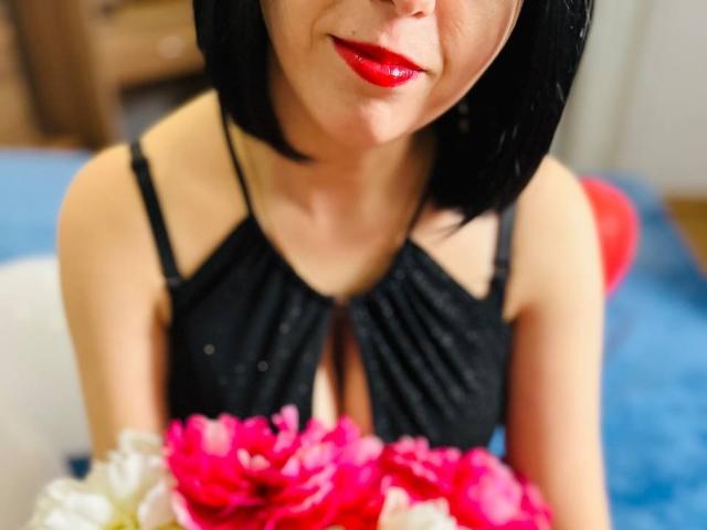 Janel with black bob, red lipstick, black dress, holding pink and white flowers, showing her tits, blue background, red balloon blurred in the background.