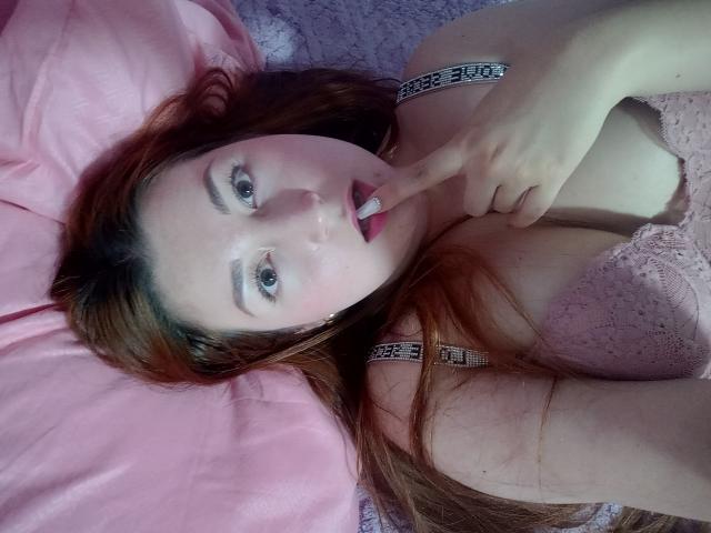 ReginaLov with red hair, pink lace bra, and finger in mouth, lying on pink bed, showing her tits and slightly open mouth, looking at camera with blue eyes.