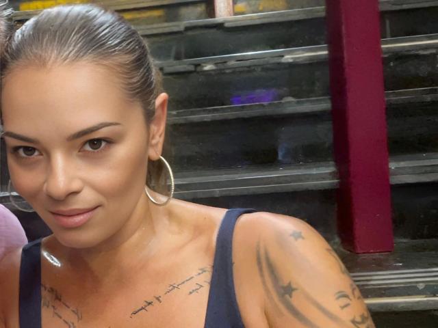 Brownsugarheaven-hot with slicked-back brown hair, wearing large hoop earrings, blue tank top, and tattoos on her shoulder, smiling slightly at the camera, showing her tan skin.
