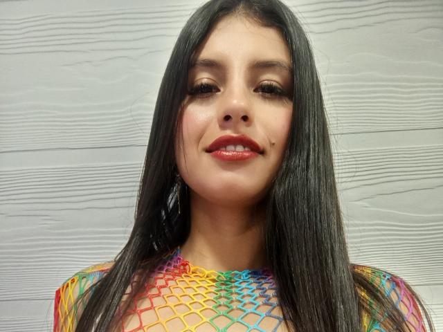 MadissonFontaine with long black hair, wearing a colorful fishnet top, has red lipstick and a slight smile, showing her shiny nose and dark eyes.