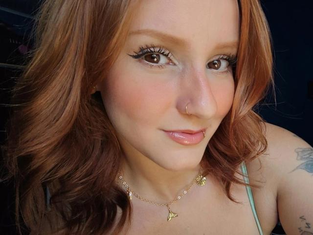 Ruivaxx has light brown wavy hair, brown eyes with black eyeliner, a nose ring, pink lips, and a gold necklace with small triangles. Her tattoo is partially visible.
