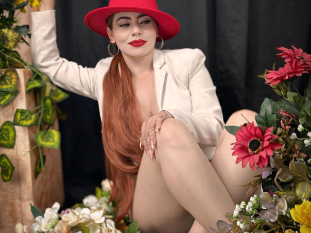 DulceCharlotteHot with long red hair, red hat, white blazer, red lipstick, sitting with knees up, surrounded by flowers, earrings, and green plant leaves, looking sexy and confident.