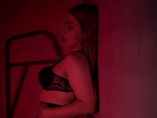 DulceCharlotte, peau rosée, portant un soutien-gorge en dentelle noire, debout de côté contre un mur, les yeux fermés, la bouche légèrement ouverte, une ombre sur le mur, éclairage rouge..