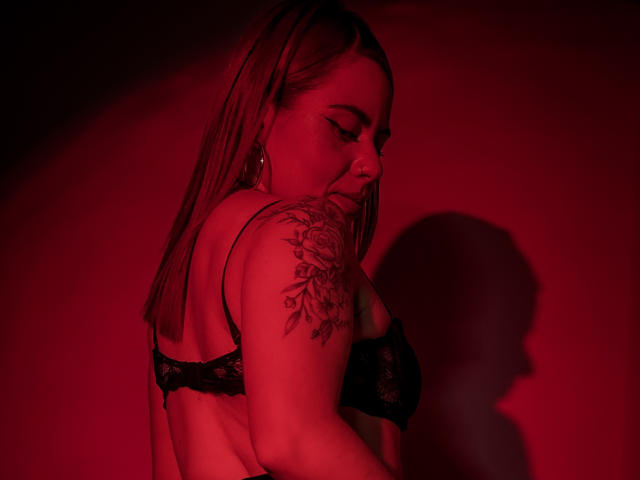 DulceCharlotteHot with red-tinted lighting, wearing black lace bra, showing tattooed shoulder, long straight brown hair, hoop earrings, and shadow on wall. Boobs visible, looking down.