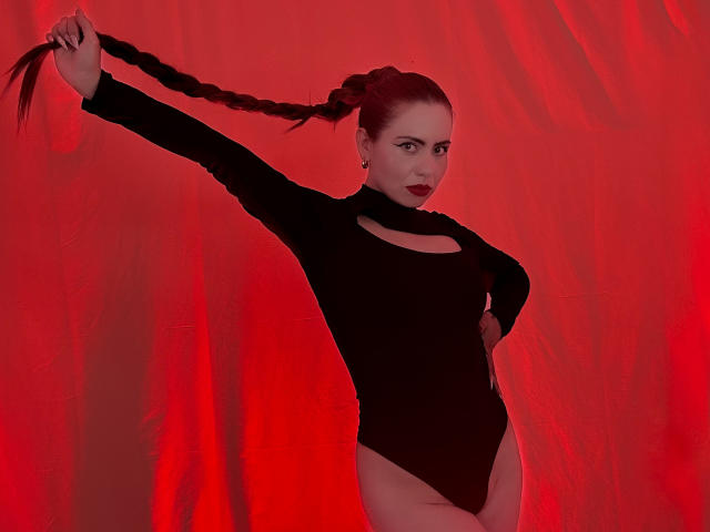 DulceCharlotteHot with long braided hair, wearing a black long-sleeve leotard, holds her hair out, red background, red-lipsticked, standing with hand on hip, showing her ass.
