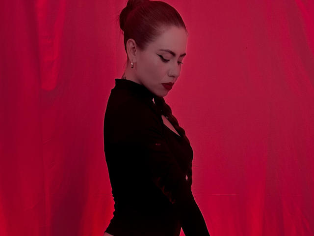 DulceCharlotteHot with red hair, black dress, and earrings, looking down with closed eyes, has a red light on her. She's got a cute ass and dark red lipstick.