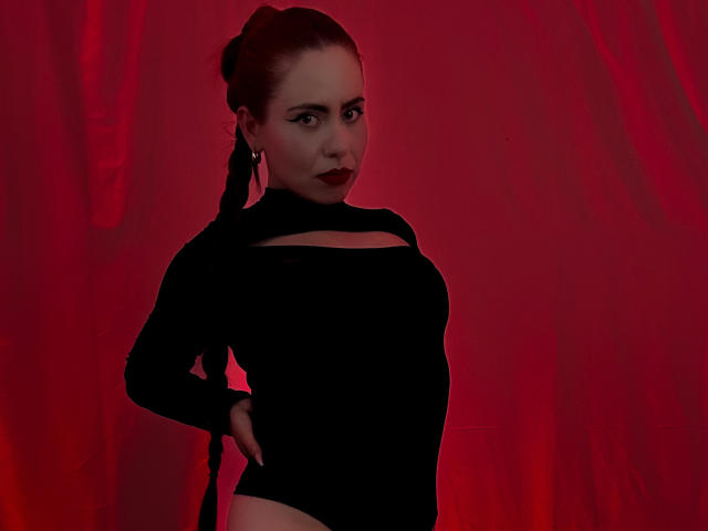 DulceCharlotteHot with dark red lipstick, black long-sleeve top, and high-cut black bottom, hand on hip, red background, braided hair, confident, sexy, looking at camera.