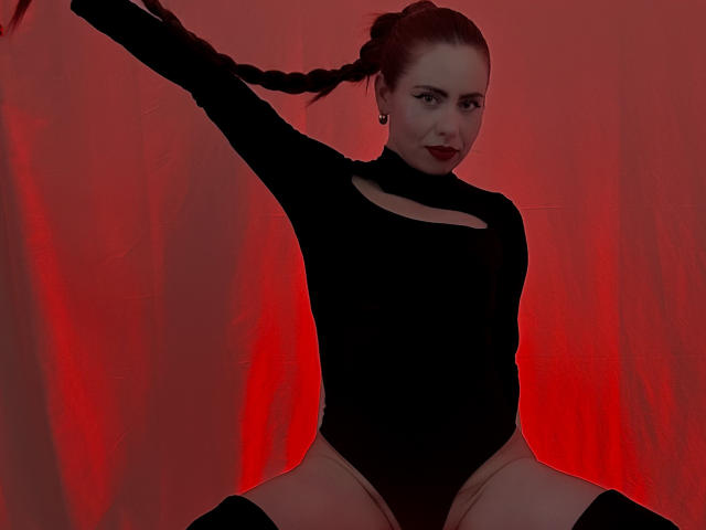 DulceCharlotteHot with braided hair, wearing a black cutout leotard, has her arms raised, showing her tits, and is sitting with her legs spread, against a red backdrop.