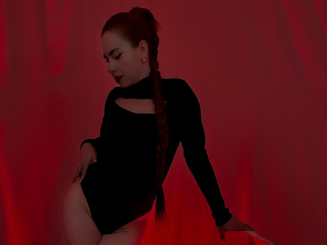 DulceCharlotteHot with a braided red ponytail, wearing a black cutout leotard, sitting back with a hand on her ass, red lighting, looking down, dark lipstick.