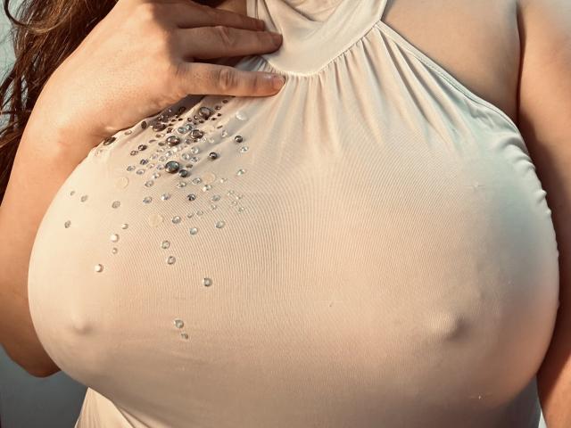BadAngel's large tits, covered by a wet, white halter-top with sparkling beads, showing nipples through the fabric. Her hand grips the top, sunlight highlighting her skin.