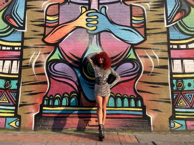 Cinthia Walker está de pie frente a grafitis coloridos, vestida con un vestido de estampado de leopardo, botas negras y cabello rizado rojo, con una mano en la cadera, mostrando su trasero..