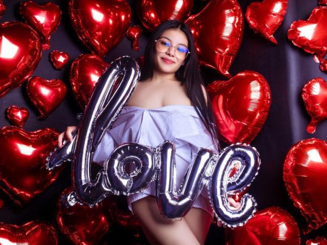Ebba with blue glasses, long dark hair, white off-shoulder dress, holding shiny "love" balloon, surrounded by red heart balloons, showing her bare ass and tits.