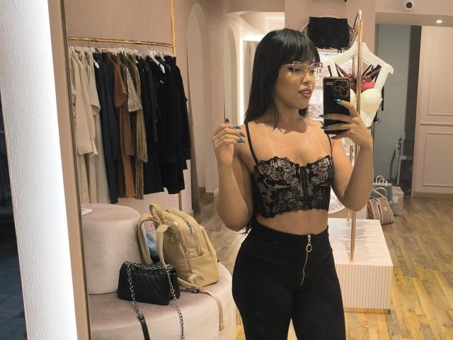 Heyly macht ein Spiegelselfie in einem Geschäft, trägt einen schwarzen Spitzen-Bralette und eine schwarze Hose mit hohem Bund und lächelt.