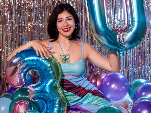 AnastasiaEvansX, with short brown hair, red lipstick, and blue-green dress, holds shiny blue number balloons, surrounded by purple and silver balloons, smiling at the camera.