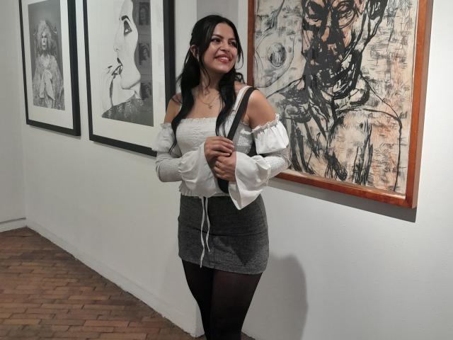 EliieRuizz with long black hair, smiling, wearing off-shoulder white blouse, gray skirt, black tights, standing in front of abstract and black-and-white framed art.