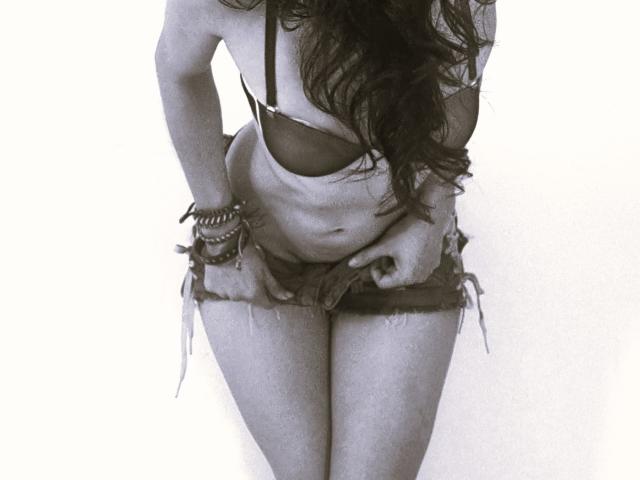 HarlyMia's tits in a black bra, wearing frayed denim shorts, hand on her crotch, long wavy hair, dark skin, multiple bracelets, standing with legs together, black-and-white photo.