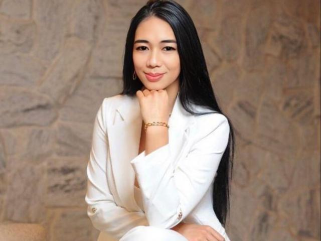 Orka with long black hair, wearing a white jacket, smiling softly, hands near face, gold bracelet on right wrist, cute, attractive, sitting, relaxed, confident, sexy.