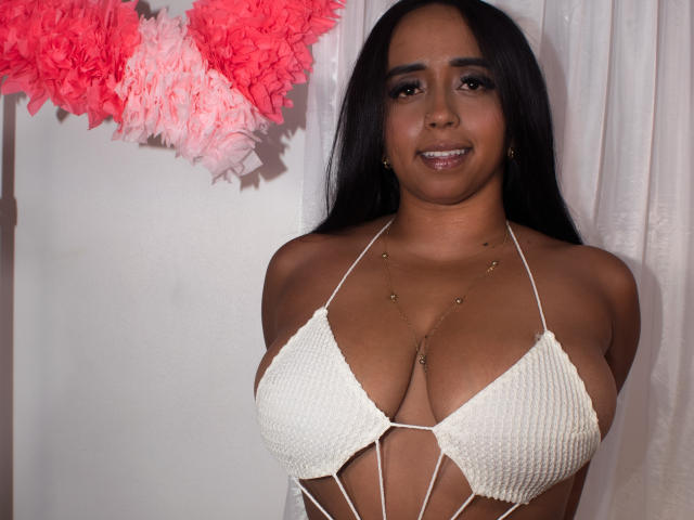 Stacy Cossio, avec sa peau bronzée, ses cheveux noirs et ses yeux marron, porte un haut de bikini blanc à fines bretelles croisées, qui met en valeur sa poitrine généreuse et son petit collier en or..