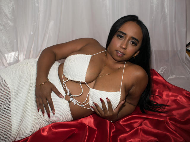 Stacy Cossio, à la peau foncée, avec de longs cheveux noirs, portant un haut de bikini blanc en tricot qui révèle sa poitrine généreuse et une jupe blanche en tricot, allongée sur du satin rouge, regardant l'appareil photo..