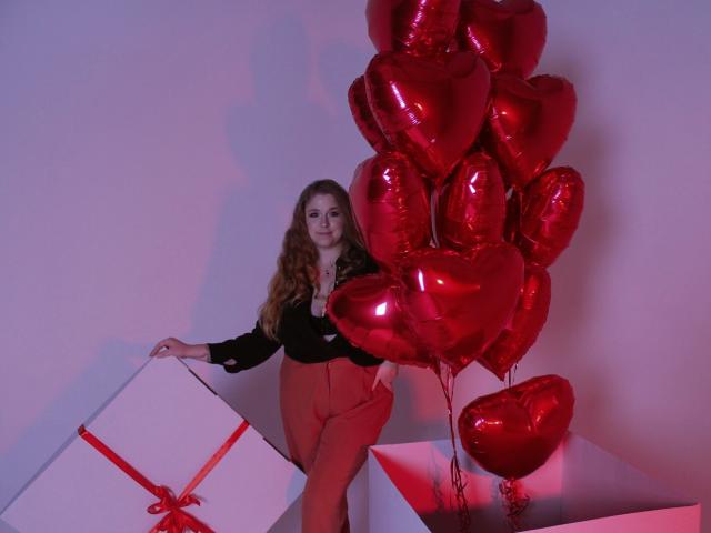 Jessika Daniels, con cabello castaño ondulado, blusa negra, pantalones rojos, sosteniendo una caja blanca con una cinta roja, de pie junto a globos en forma de corazón rojos, en una habitación iluminada con luz púrpura..