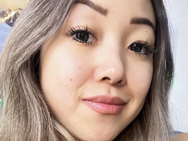 RenyLime's face with light brown, wavy hair, green eyes, and long, dark eyelashes. She has pink, glossy lips and a subtle smile. Her skin is fair with small freckles.