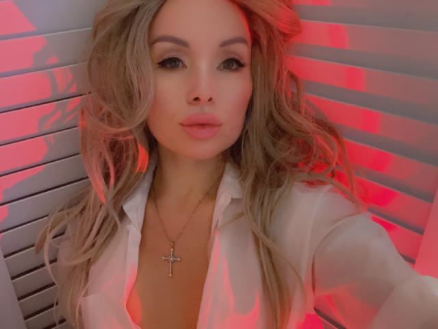 AvaGreenx, con cabello rubio ondulado, vestida con una camisa blanca, un collar con una cruz, iluminación roja y labios rosados fruncidos, mirando directamente a la cámara..