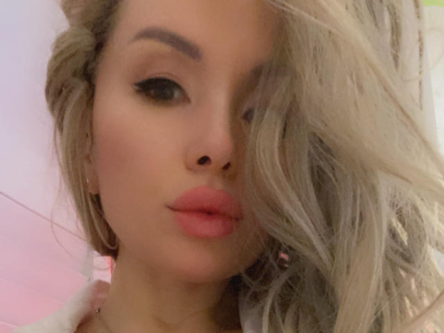 AvaGreenx has blonde hair, full pink lips, and subtle makeup with black eyeliner. Her wavy hair covers part of her face, and she wears small hoop earrings.