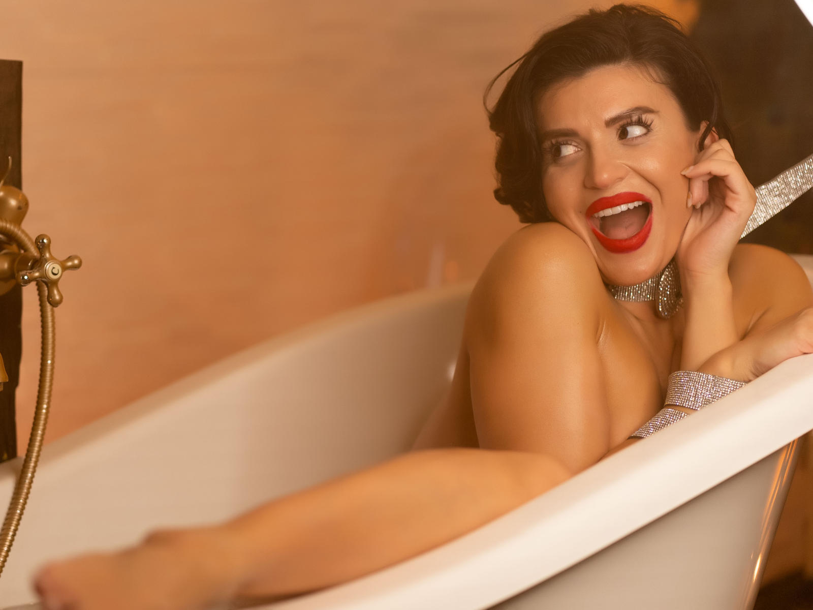 OnlyNicolle in a white bathtub, smiling with red lipstick, holding her ear, showing her bare tits and shiny necklace, with dark hair and a gold faucet.
