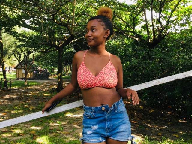 BelleXShara with brown skin, small tits in a pink patterned bra, and a yellow bun, leans on a white fence in a forest, wearing a blue, ripped, denim skirt.