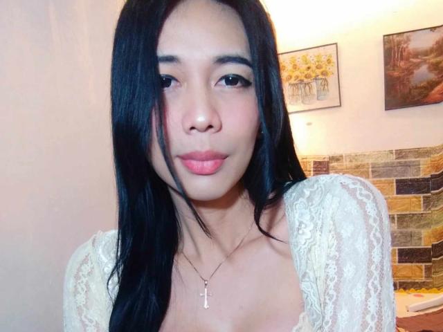 Orka has long black hair, light brown skin, and is wearing a lace top, with pink lips and a cross necklace, showing her tits slightly in the selfie.