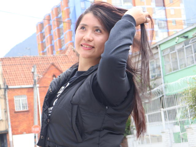 AshleyWalkerI with long brown hair, smiling, wearing a black jacket, hand in hair, exposing her tits, standing outside, city background, clear day, casual look, cute.