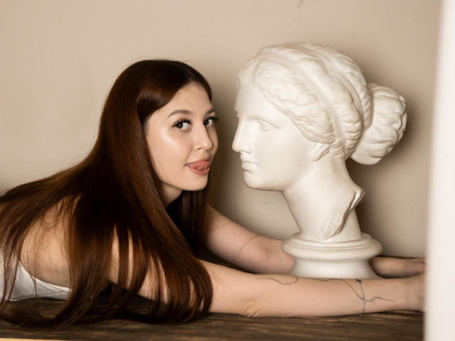 EllyCat with long brown hair, lying next to a white classical bust, pouting slightly with a tattoo on her arm, in a soft, neutral-colored setting.