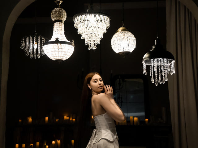 EllyCat with long brown hair, wearing a strapless white dress, stands under four glowing crystal chandeliers, hands near face, in a dimly lit room with candles in the background.