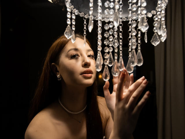 EllyCat has long brown hair, light skin, and is looking up at a crystal chandelier with droplet-shaped ornaments, her hands gently touching the sparkling hangings. She's wearing a pearl necklace.