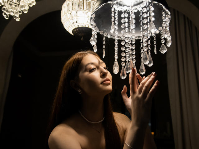EllyCat with brown hair, fair skin, and pearl necklace, eyes closed, hands up, under a hanging crystal chandelier, looking at dangling crystals, soft light on her face.