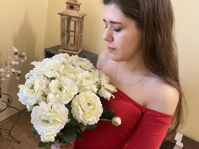 JoannaSia with long brown hair, wearing red off-shoulder top, holding white roses, looking down, silver necklace, wooden lantern in background, soft yellow wall, wooden floor.