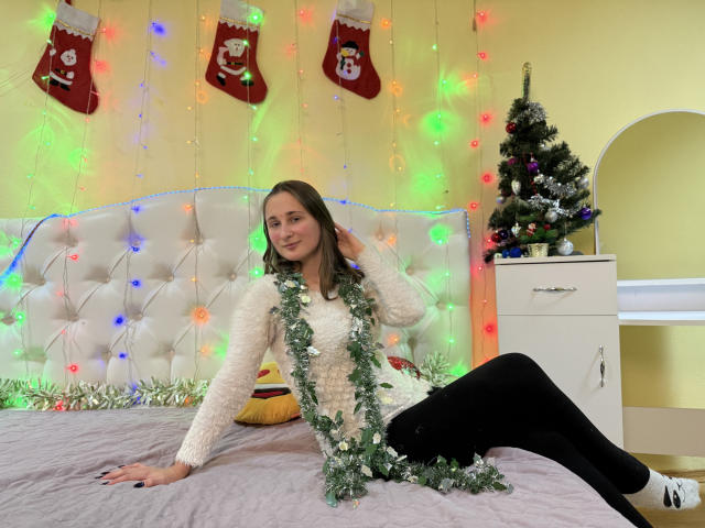 MichelleHotX69 with long brown hair, white fuzzy sweater, black pants, green garland, sitting on bed, smiling, Christmas tree on right, colorful lights, yellow wall, and red stockings.