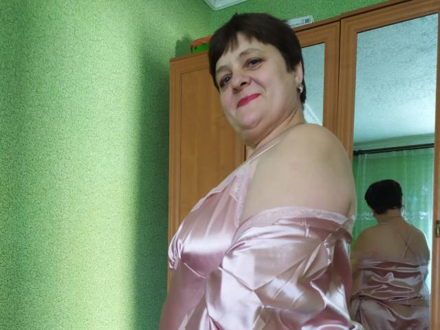 ElzaJewel, a chubby woman with short brown hair, pink satin dress, bare shoulders, smiling, reflected in mirror, green textured wall, wooden wardrobe, pink lipstick.